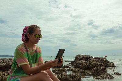 Woman using digital tablet while sitting at beach against sky