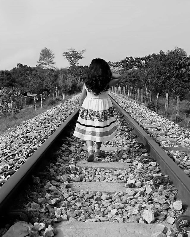 railroad track, full length, lifestyles, railing, rear view, leisure activity, tree, standing, the way forward, rail transportation, casual clothing, walking, diminishing perspective, clear sky, day, stone - object, sky, connection