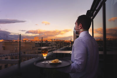 Waiter wearing mask with tray standing at outdoor restaurant during sunset