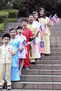 Portrait of cute friends in traditional clothing holding malaysia flags while standing on steps