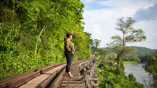 Side view of woman standing on railroad track by trees against sky