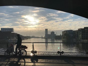 Man with bicycle standing by railing of bridge in city against sky