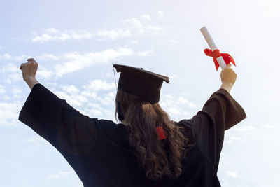 Rear view of woman in graduation gown standing with arms outstretched against sky