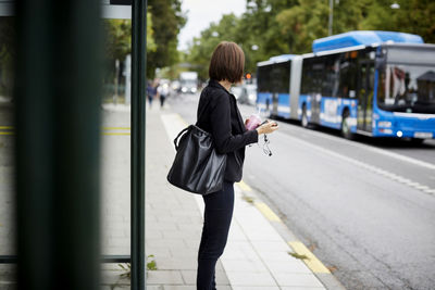 Side view of businesswoman standing on sidewalk waiting at bus stop in city