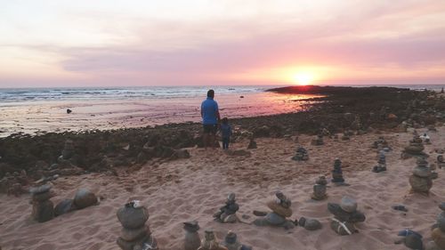 Rear view of father standing with daughter by stacked stones at beach during sunset