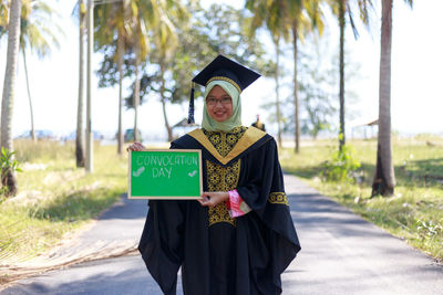Portrait of young woman in graduation gown on road with writing slate