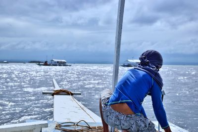 Rear view of man crouching on boat in sea against cloudy sky