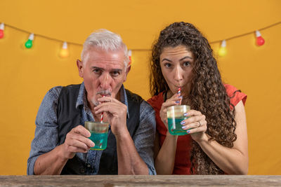 Father and daughter drinking a blue cocktail sitting on yellow background - studio shot