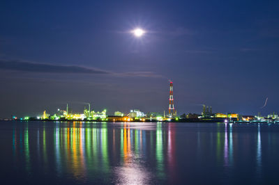 Illuminated factory by sea against sky at night