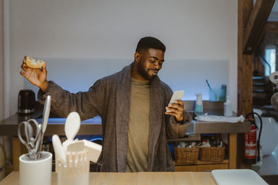 Happy man holding doughnut and using internet on smart phone at home