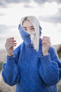 Joyful cheerful young blonde female in blue knitted sweater looking at camera and laughing while standing against blurred background of sea coast