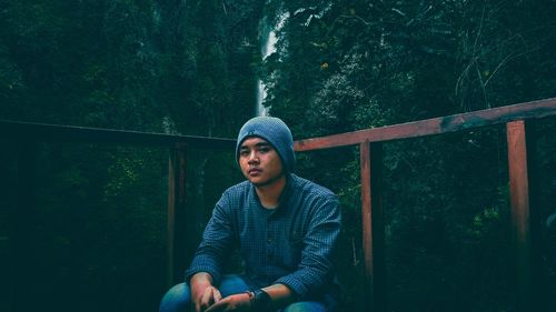 Young man looking away while sitting on railing in forest