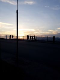 Silhouette people on street by sea against sky during sunset