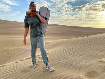 Full length of woman holding sandboard at desert during sunset