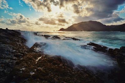 Waves splashing on rocky shore against cloudy sky during sunset