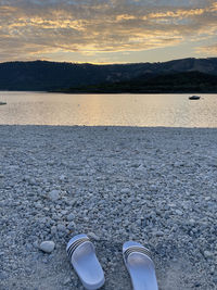 Low section of shoes by lake against sky during sunset