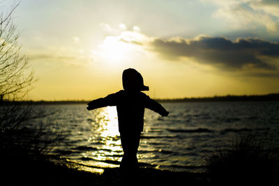 Silhouette boy standing in sea against sky during sunset