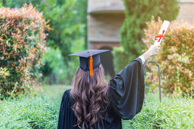 Rear view of woman in graduation gown holding certificate while standing by plants