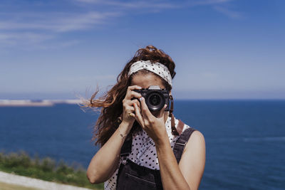 Portrait of young woman photographing sea against sky