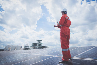 Low angle view of worker wearing reflective clothing while standing on solar panel against sky