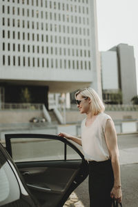 Smiling woman holding car door while standing against building in city