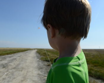 Boy standing on road amidst grassy field against sky during sunny day