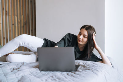 Beautiful smiling woman student freelancer with dark long hair in casual using laptop on bed at home