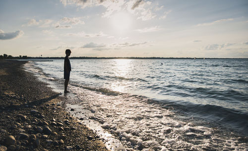 Full length of silhouette boy standing on shore at beach against sky during sunset