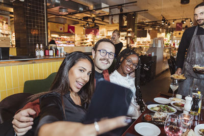 Cheerful woman taking selfie through smart phone with male and female friends in restaurant