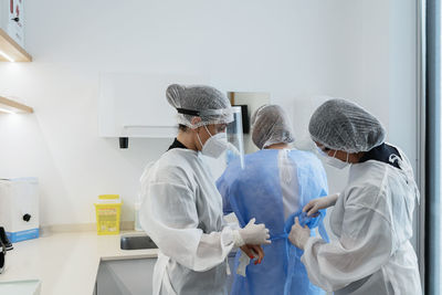 Female medical workers in protective masks and face shields helping each other to get dressed while preparing for medical procedure in hospital