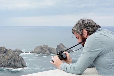 Side view of man with camera leaning on railing against cloudy sky