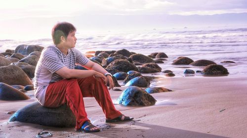 Man looking away while sitting on rock at beach against sky