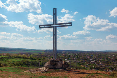 Large metal cross standing on hilltop, overlooking a serene village and expansive fields 