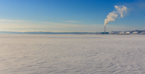 Distant view of smoke stack emitting pollution on snow covered field against sky