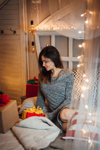 Woman looking away while sitting on illuminated lamp at home