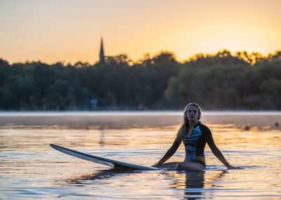 Woman sitting on surfboard at lake during sunset