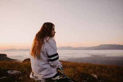 Woman looking away while sitting against sky during sunset