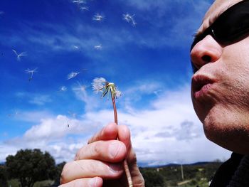 Close-up of man blowing dandelion seeds against sky