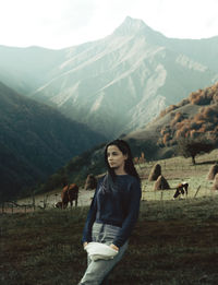 Young woman looking away while standing on grass against cows and mountains