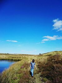 Full length of boy walking on grassy field by pond against blue sky