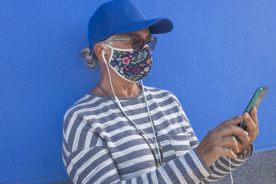 Close-up of senior woman wearing sunglasses using smart phone standing against blue wall