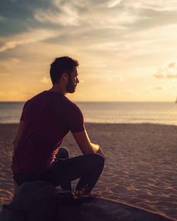 Man sitting on beach against sky during sunset