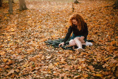 Happy young woman sitting on field with dry leaves during autumn