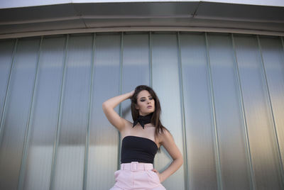 Young woman in elegant outfit looking at camera while standing near glass wall in modern building