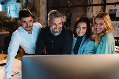 Joyful team of men and women looking at the pc screen and laughing while standing at the office