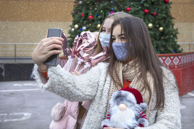 Women wearing mask photographing with mobile phone against christmas tree in winter