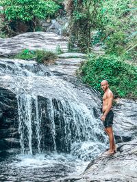 Full length of shirtless man standing by waterfall in forest