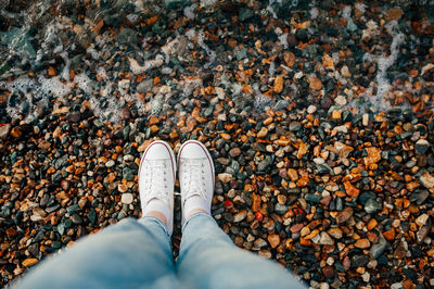 Slender legs in blue jeans and white stylish sneakers on rocky beach and wave rolls over your feet.