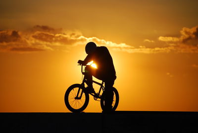 Silhouette man riding bicycle against orange sky