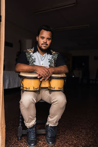 Full body of black male musician in casual clothes sitting on chair with drums while smiling and looking at camera